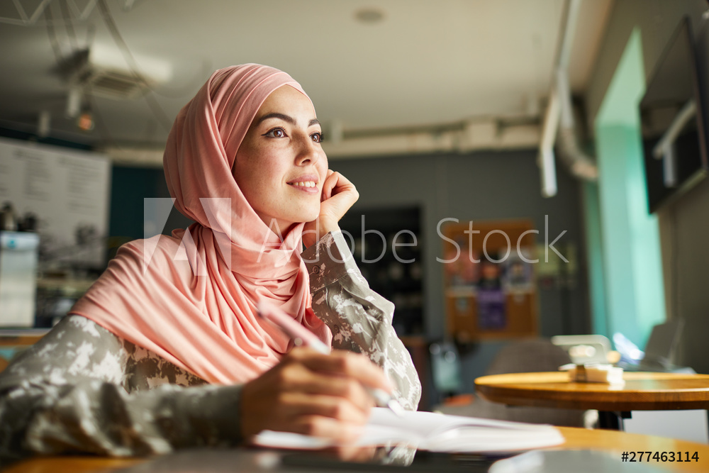 Pretty positive young woman in hijab writing her dreams in diary when sitting at table in cafe