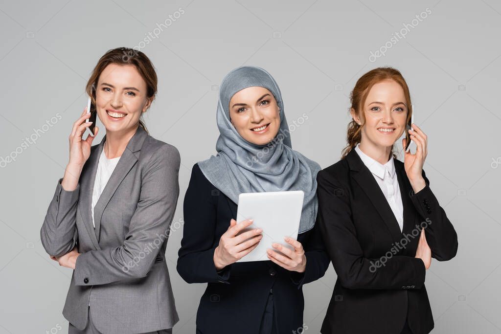 Arabian businesswoman holding digital tablet near colleagues talking on smartphones isolated on grey