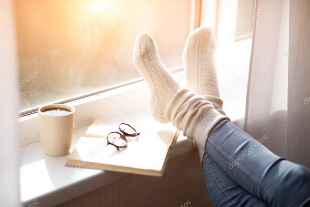 Cropped view of woman putting her feet in warm comfy socks on windowsill with book and coffee, closeup