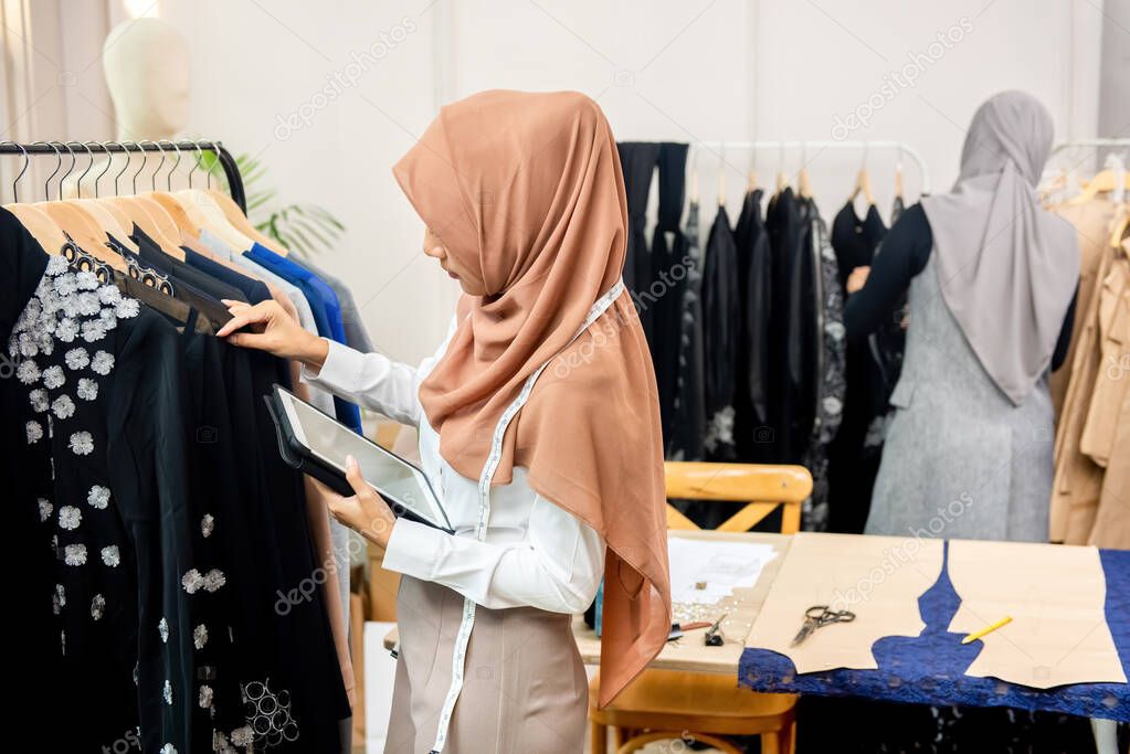 Woman muslim designer holding a tablet while checking hanged clothes in a garment boutique shop
