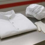 Set perlengkapan seragam kerja ruang bersih terdiri dari jas ESD, topi pelindung, dan sarung tangan lateks yang tertata rapi di atas meja stainless, menampilkan konsep higienis dan profesional dalam lingkungan cleanroom.