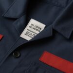 Label informasi seragam industri pada pakaian kerja berwarna navy dengan aksen merah, menonjolkan detail penting dalam standar audit dan keamanan produksi.
